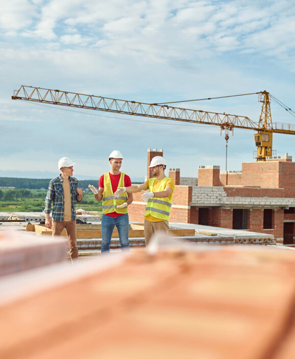 Discussion. Men in protective helmet discussing idea in positive mood standing at top of unfinished house against background of construction crane and sky on fine day